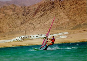 Surfen auf dem Meer in Ufernähe (50m), im Hintergrund die Berge der Sinaiwüste und einige weiße Gebäude einer Hotelanlage, die wie eine Ortschaft wirkt